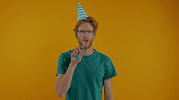 sad redhead man in party cap blowing in party horn isolated on yellow - Video