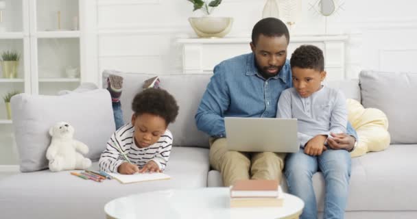 Onnellinen nuori afroamerikkalainen isä viettää aikaa pienten lasten kanssa sohvalla kotona. Isä näytti jotain kannettavalla tietokoneella pienelle pojalle. Daugther väritys kuva tai piirustus. Lapset isän kanssa. - Materiaali, video