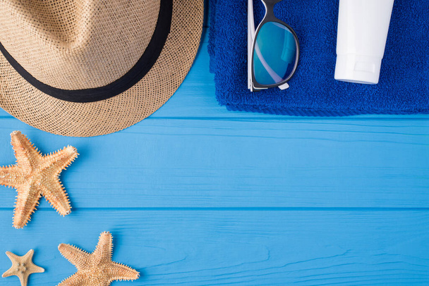 Summertime vacation time concept. Top above overhead view close-up photo of starfish towel sunglasses sunhat and sunscreen isolated on blue wooden background with copyspace - Photo, Image