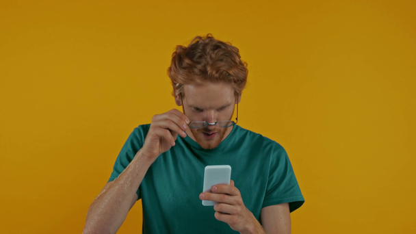 překvapený zrzka muž v brýlích pomocí smartphone izolované na žluté - Záběry, video