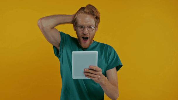 šokovaný zrzka muž v brýlích pomocí digitální tablety izolované na žluté - Záběry, video