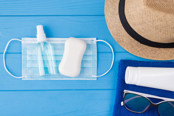 Quarantined summertime concept. Top above overhead view close-up photo of towel sunglasses sunscreen mask sanitizer sopa and sunhat isolated on blue wooden background with copyspace - Photo, Image