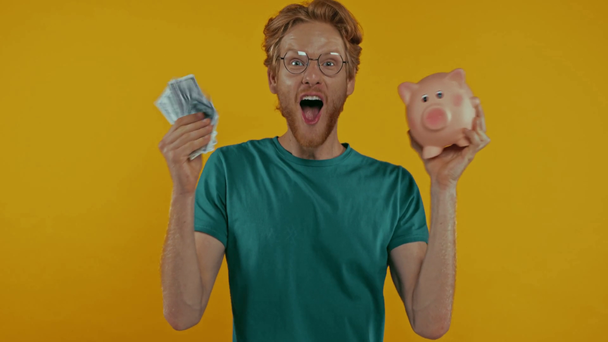 boldog vörös hajú férfi rázza malac bank és készpénz elszigetelt sárga - Felvétel, videó