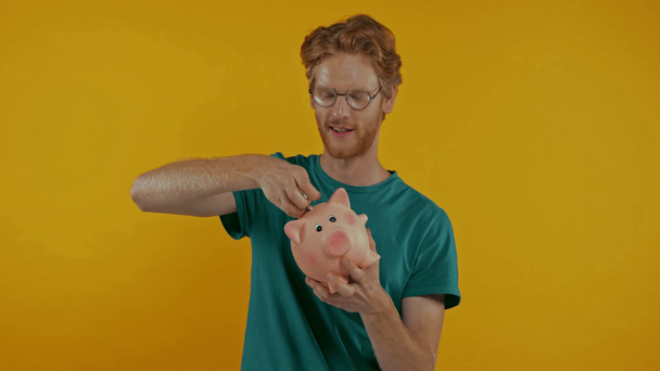 happy redhead man putting dollar in piggy bank isolated on yellow - Video, Çekim