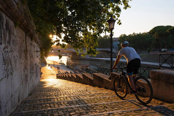Camine por el Tevere, al atardecer con el sol en un día de verano cerca de la isla del Tíber. Un ciclista camina por la escalera inundada por los rayos del sol que se está poniendo en el Tevere. Roma, Italia
. - Foto, imagen