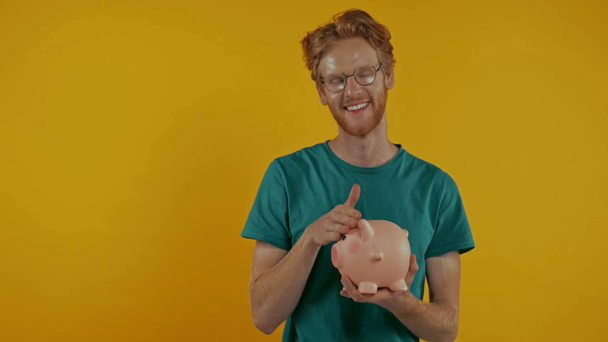 szczęśliwy ruda człowiek wprowadzenie monety w świnka bank odizolowany na żółty - Materiał filmowy, wideo