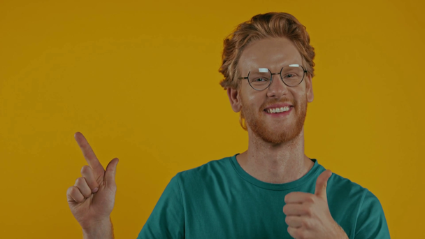 cheerful redhead man showing thumb up and pointing aside isolated on yellow - Footage, Video