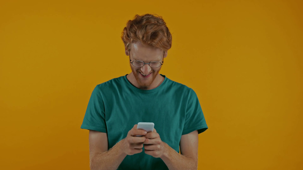 gelukkig roodharige man met bril wijzend naar smartphone geïsoleerd op geel - Video