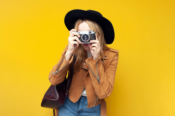 fiatal lány őszi ruhák egy hátizsák tartja a kamerát, és készít egy fényképet egy sárga elszigetelt háttér - Fotó, kép