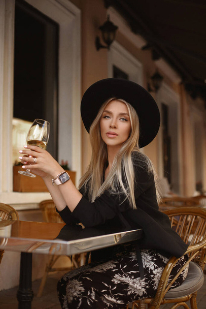 A gorgeous young woman in a black hat enjoying a glass of wine and looking in camera at the cafe table on the street in a summer evening - Foto, Bild