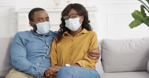 Portrait of African American young couple in medical masks siting on couch at home. Coronavirus concept. Man and woman looking at camera on sofa in hugs in living room on lockdown. Male and female. - Footage, Video
