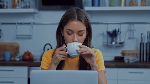 hübsche junge Frau trinkt Kaffee in der Nähe von Laptop  - Filmmaterial, Video