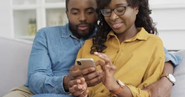 若いアフリカ系アメリカ人の陽気なカップルがソファに座ってスマートフォンの画面で何かを見ています。家で。携帯電話を使用してハンサムな男と美しい女性は、タップしてスクロールします。室内. - 映像、動画