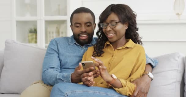 アフリカ系アメリカ人の若いハンサムな男は、かなりの女性の笑顔と抱擁。幸せなカップルソファに座って、自宅で携帯電話で何かを見て。スマホを使った彼女と彼氏. - 映像、動画