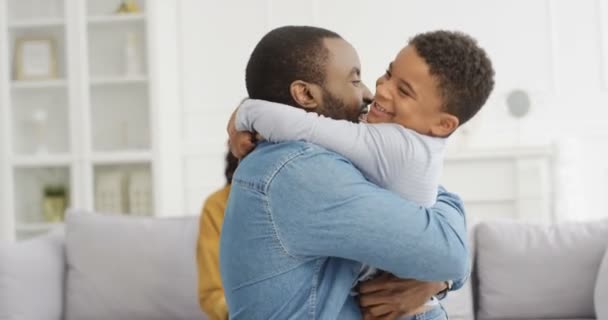 Feliz hombre y mujer afroamericanos alegres abrazando a su pequeño hijo lindo y riendo en casa en el sofá. Padres alegres con un niño pequeño en el salón. Madre y padre sonriendo y abrazando al niño. - Metraje, vídeo