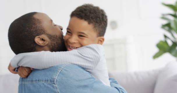 Feliz hombre afroamericano alegre abrazando a su pequeño hijo lindo y riéndose en casa en el sofá. Padres alegres con niños pequeños en el salón. Padre joven sonriendo y abrazando al niño en el interior. Generaciones - Metraje, vídeo
