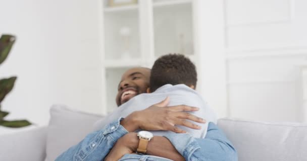 Счастливый веселый афроамериканец обнимает своего маленького милого сына и смеется над домом на диване. Радостный родитель с маленьким ребенком в гостиной. Молодой отец улыбается и обнимает ребенка внутри. Задняя часть. - Кадры, видео