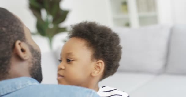 A boldog afro-amerikai férfi háta mögött ölelgeti aranyos kislányát, és otthon nevet a kanapén. Vidám szülő kisgyerekkel a nappaliban. Visszapillantás az apa mosolygós és átölelő gyermek - Felvétel, videó