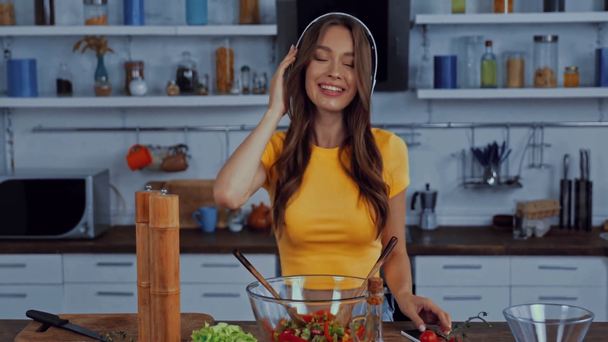 onnellinen nainen langattomissa kuulokkeissa tanssia ja sekoittamalla salaattia - Materiaali, video