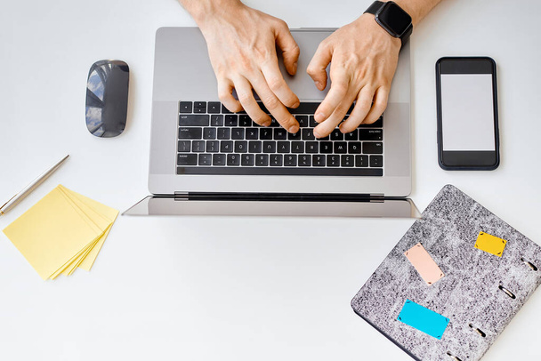 Een man typt tekst, werkt achter een laptop, naast een telefoon met een witte kraan - Foto, afbeelding