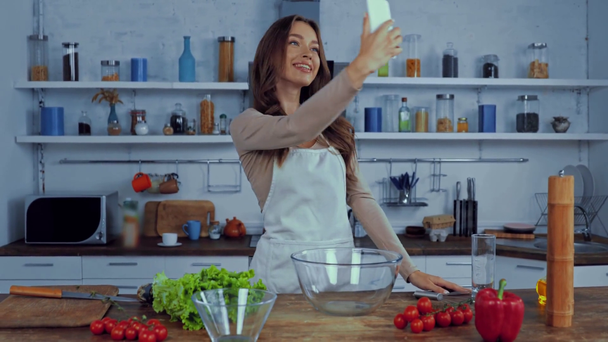 Glückliche Frau in Schürze macht Selfie nahe Zutaten auf Tisch - Filmmaterial, Video