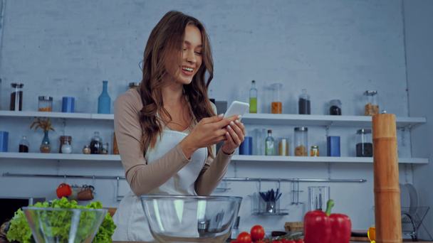 felice donna in grembiule utilizzando smartphone vicino ingredienti sulla tavola  - Filmati, video