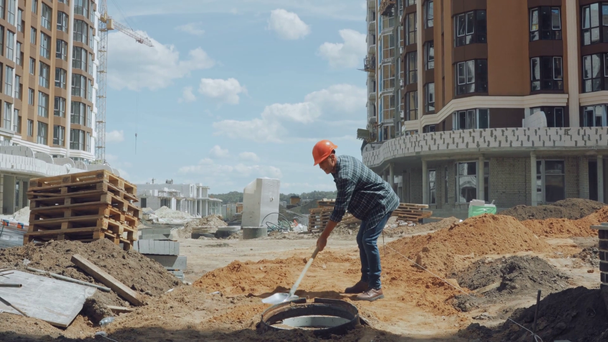 Builder in hard hat digging ground with shovel on construction site - Materiaali, video