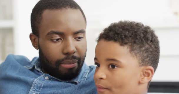 Κοντινό πλάνο του Αφροαμερικανού μικρού αγοριού που κάθεται με τον όμορφο πατέρα του στο σπίτι, τρώγοντας ποπ κορν και βλέποντας τηλεόραση. Εσωτερικά. Ο μπαμπάς και ο γιος περνούν χρόνο μαζί.. - Πλάνα, βίντεο