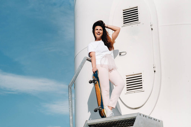 A girl in white clothes with a skate in her hands is photographed near large wind turbines in a field with trees.Modern woman with a Board for riding in a field with windmills. - Zdjęcie, obraz