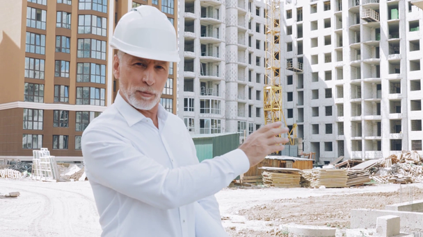 Engineer with blueprint talking at camera while pointing at construction site - Footage, Video