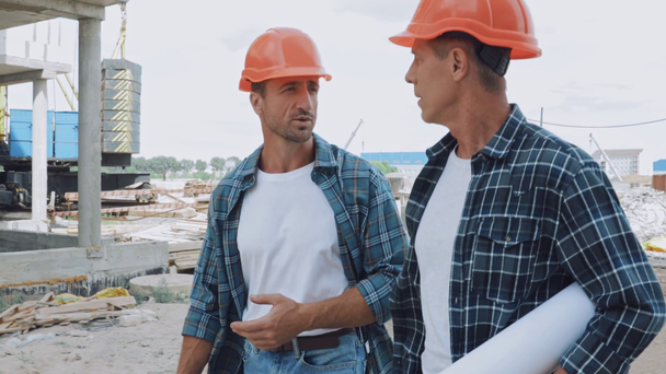Építők kemény kalapban tervrajzzal beszél, miközben sétál az építési területen - Felvétel, videó