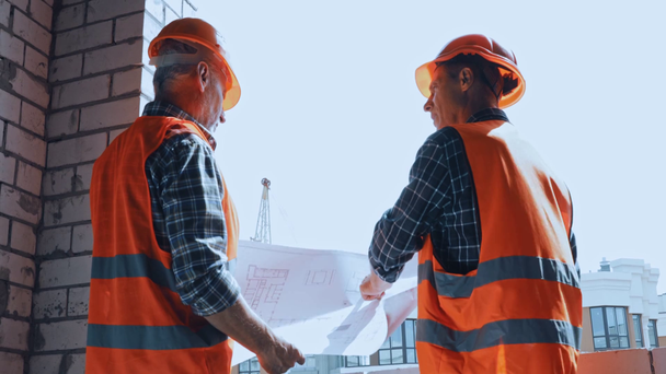 Builders in hard hats discussing blueprint on construction site - Metraje, vídeo