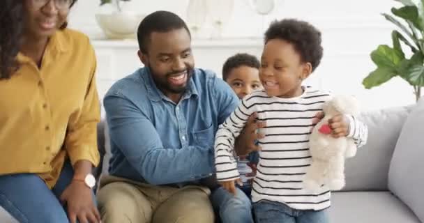 若い幸せなアフリカ系アメリカ人の両親の肖像画ソファの上にカメラに陽気に笑みを浮かべて座っている。母と父と一緒に楽しい子供たちは笑って、リビングルームでソファで楽しんでいます. - 映像、動画