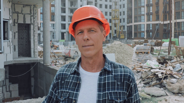 Jóképű építész kemény kalapban mosolyog a kamera előtt az építkezésen - Felvétel, videó