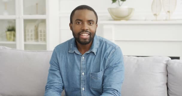 Portret van knappe jonge Afro-Amerikaanse man blogger zittend op de bank in de woonkamer en pratend met camera. Een mannelijke vlogger die iets vertelt en uitlegt. Videoblog. Vlog van thuis. - Video