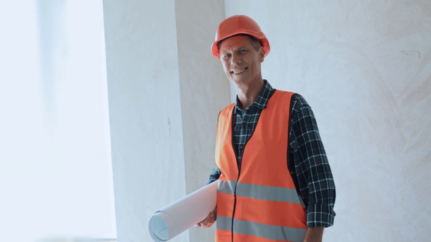 Uśmiechnięty budowniczy trzymający plan i patrzący na kamerę na placu budowy - Materiał filmowy, wideo