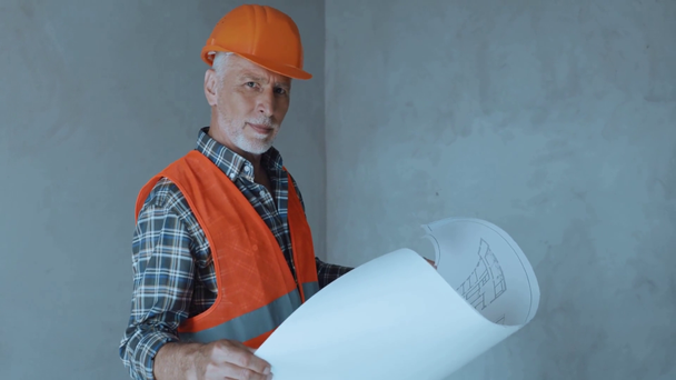 Stavitel drží plán a usmívá se na kameru na staveništi - Záběry, video