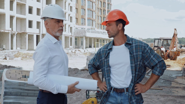 Инженер с чертежом и строитель в каске разговаривает на строительной площадке - Кадры, видео