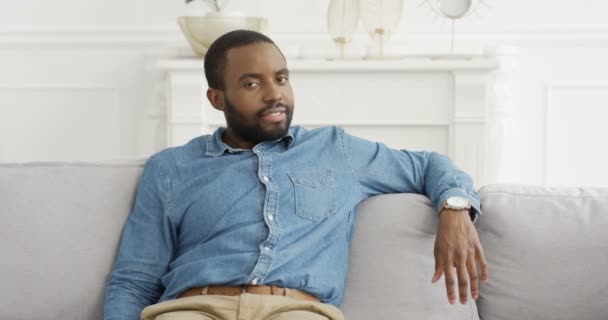 Retrato de belo jovem afro-americano de camisa jeans azul sentado no sofá e sorrindo para a câmera. Bom aspecto masculino com sorriso no sofá em casa. - Filmagem, Vídeo