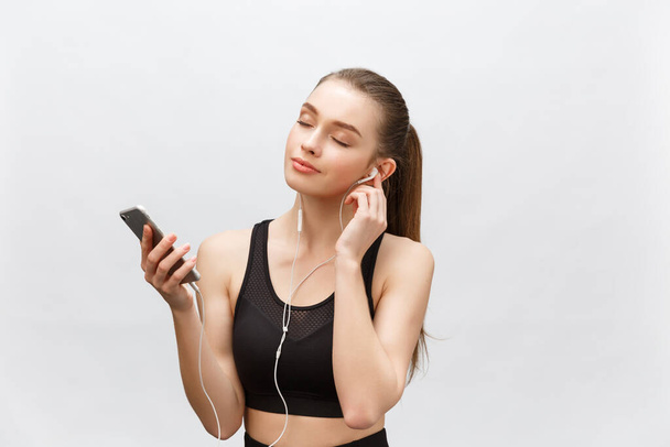 Isolated shot of young sportswoman has healthy athleteic body, listens music with headphones, holds smart phone. Isolated over grey background. - Valokuva, kuva