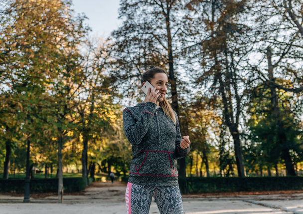 Junge blonde Frau im Sportanzug lächelt, während sie an einem Herbsttag in einem Park mit ihrem Smartphone spricht - Foto, Bild
