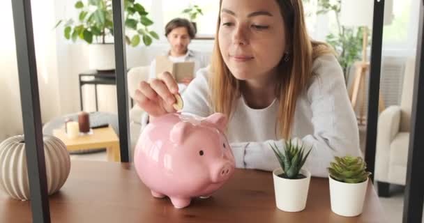 Vrouw geld besparen in spaarvarken bank en ontspannen met man lezen boek op de bank thuis - Video