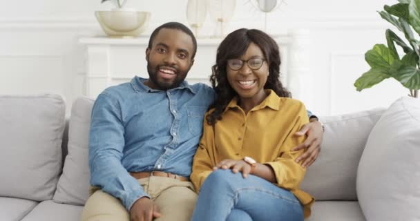 Portret młodej afroamerykańskiej szczęśliwej pary siedzącej w uściskach na kanapie i rozmawiającej z kamerą jak blogerzy. Ładna kobieta i przystojny mężczyzna uśmiechający się radośnie i mówiący coś. Wideo w domu. - Materiał filmowy, wideo
