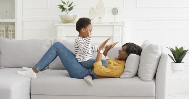 Afrikkalainen amerikkalainen onnellinen kaunis äiti pelaa kädet peli pieni söpö tytär sohvalla kotona. Äiti ja lapsi viettävät aikaa yhdessä sohvalla olohuoneessa. Äitiyden ja lapsuuden käsite. - Materiaali, video