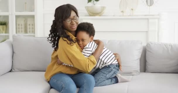 Afro-Amerikaanse jonge mooie vrouw met een bril zittend op de bank thuis en knuffelend haar schattige kleine dochter. Klein meisje omarmt moeder op de bank in de woonkamer. Moederliefde. Gelukkige veilige jeugd. - Video