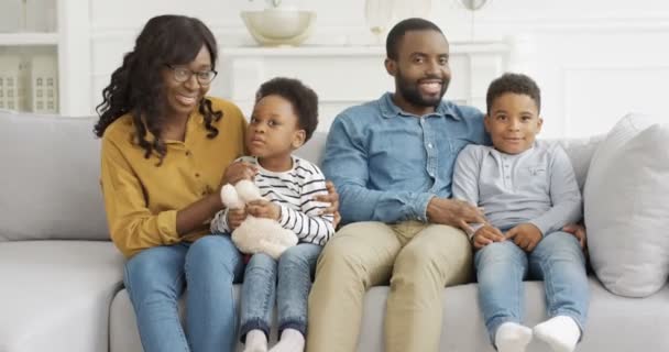 Portret van een vrolijke Afro-Amerikaanse familie met twee kinderen die op de bank zitten en vrolijk glimlachen voor de camera. Kleine leuke jongen en meisje met moeder en vader thuis. Ouders met kleine kinderen. - Video
