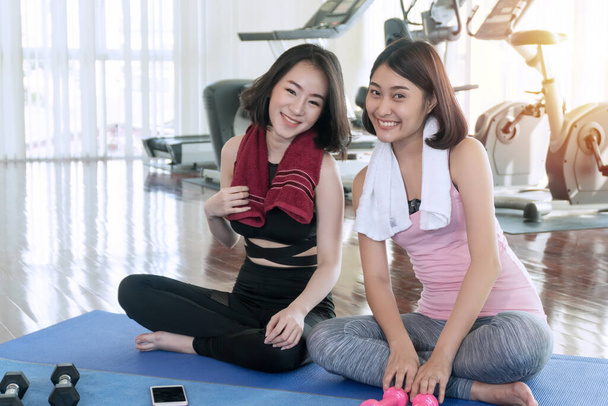 Healthy smile young woman slim body wear sportwear with towel sitting on mat yoga togetherness relaxing while successful fitness class after training at gym. Wellness exercising concept. - Photo, Image