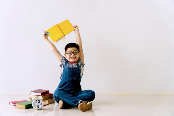 Μικρό χαριτωμένο αγόρι φορούν γυαλιά που κάθονται στο πάτωμα με στοίβα από βιβλία, μολύβι και ρολόι, ενώ κοιτάζοντας κάμερα με χώρο αντίγραφο. Ο σχολιαρόπαιδο κρατούσε ένα βιβλίο στο κεφάλι του. Επιστροφή στο σχολείο έννοια. - Φωτογραφία, εικόνα