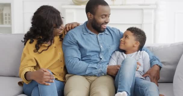 Portret van een gelukkige Afro-Amerikaanse familie met een schattig klein kind. Kleine jongen zittend op de bank in de woonkamer met ouders, elkaar aankijkend en glimlachend naar de camera. Moeder en vader met zoon thuis. - Video