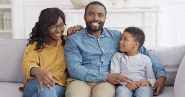 Portret van jonge Afro-Amerikaanse gelukkige familie met schattige kleine zoon. Kleine jongen zit op de bank in de woonkamer met ouders en glimlacht vrolijk. Moeder en vader met kind op de bank thuis. - Video